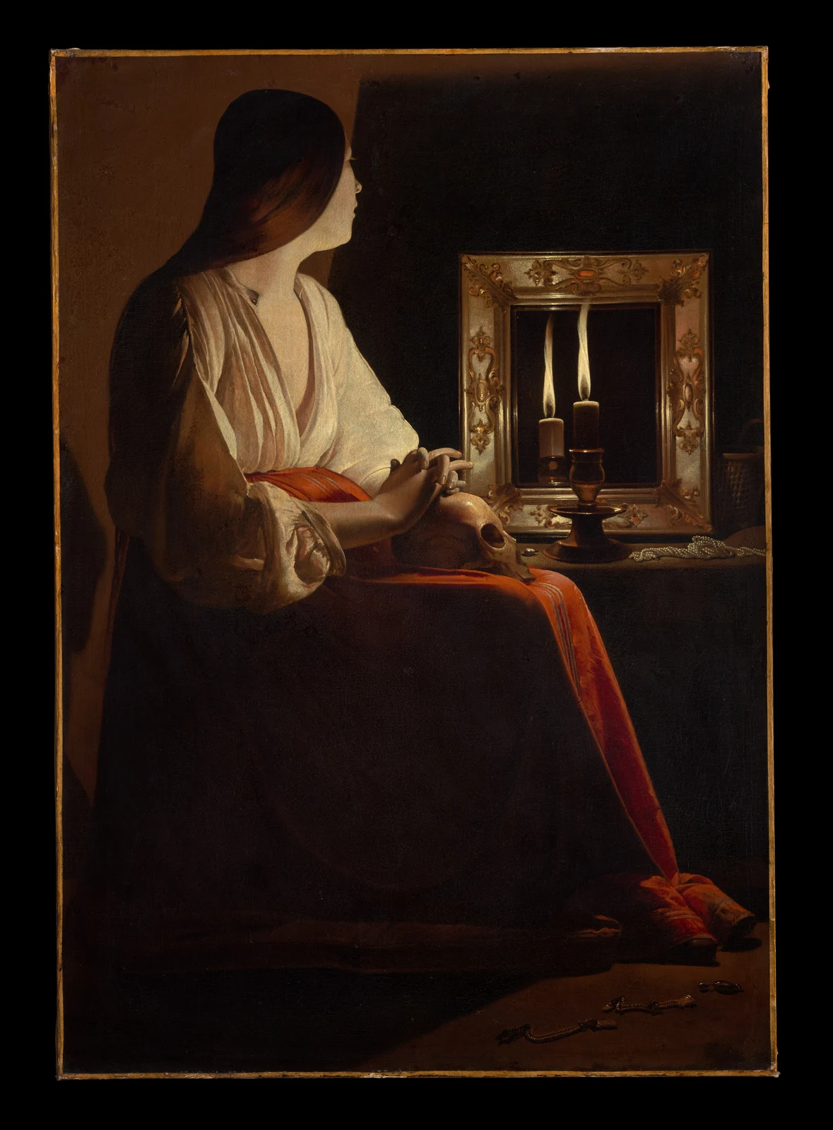A woman sits in a dark room beside a candle and its reflection in a mirror, her hand resting near a skull on her lap.