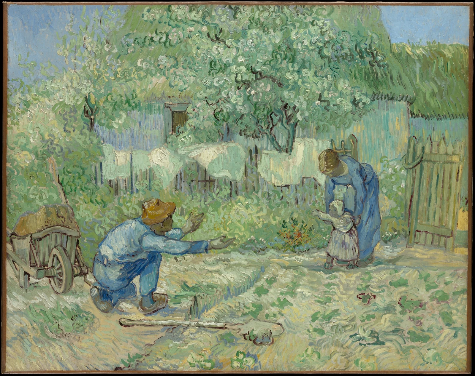 In a garden, one adult steadies a standing child while another kneels with open arms, waiting for the child to walk across.
